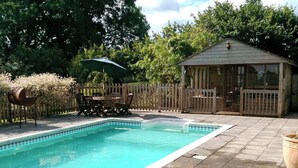 Una piscina rectangular con borde de baldosas, rodeada por una valla de madera y una estructura de madera con ventanas.
