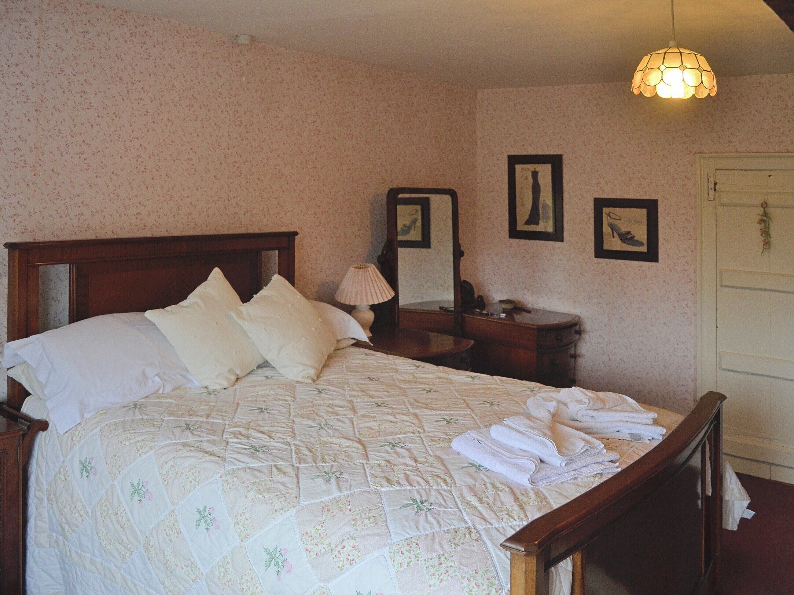 A neatly made bed with pillows, a bedside lamp, and a dresser with a mirror. There are framed pictures on the wall and a door with a decorative wreath.