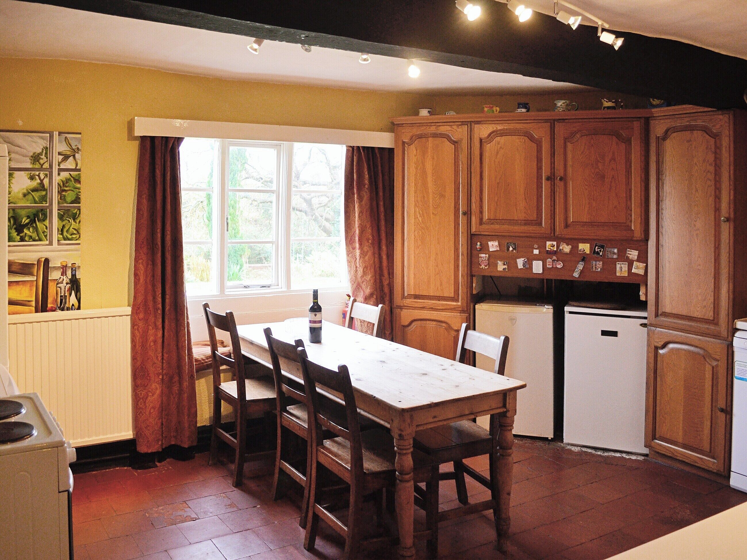 A kitchen with wooden cabinets, a dining table, chairs, a refrigerator, and a window with curtains.