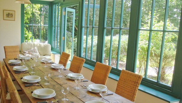 Un comedor con una larga mesa de madera dispuesta para una comida, rodeada de sillas de mimbre y grandes ventanales que ofrecen vistas a la vegetación exterior.
