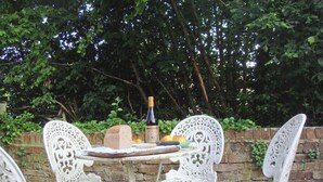 Un conjunto de mesa y sillas de hierro forjado blancas al aire libre, con un muro de ladrillo y follaje en el fondo.