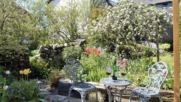 A garden patio with two metal chairs and a table, surrounded by lush greenery and flowering plants. - 1 bedroom accommodation in Lowick Bridge (Lowick Bridge)