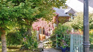 Ein Gartenweg führt zu einem Haus mit einer Holztür, umgeben von üppigem Grün und blühenden Pflanzen.