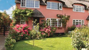 Una casa tradicional vermella amb teulada de palla, nombroses finestres i un jardí ben cuidat amb flors roses.