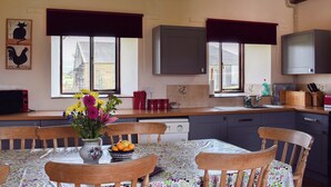 A kitchen with wooden cabinets, a dining table with chairs, and a window with a view of a house. - 3 bedroom accommodation in Brighstone (Brighstone)