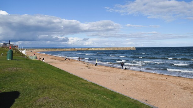 Une plage avec une zone herbeuse, un rivage sablonneux et une digue de pierres visible au loin.
