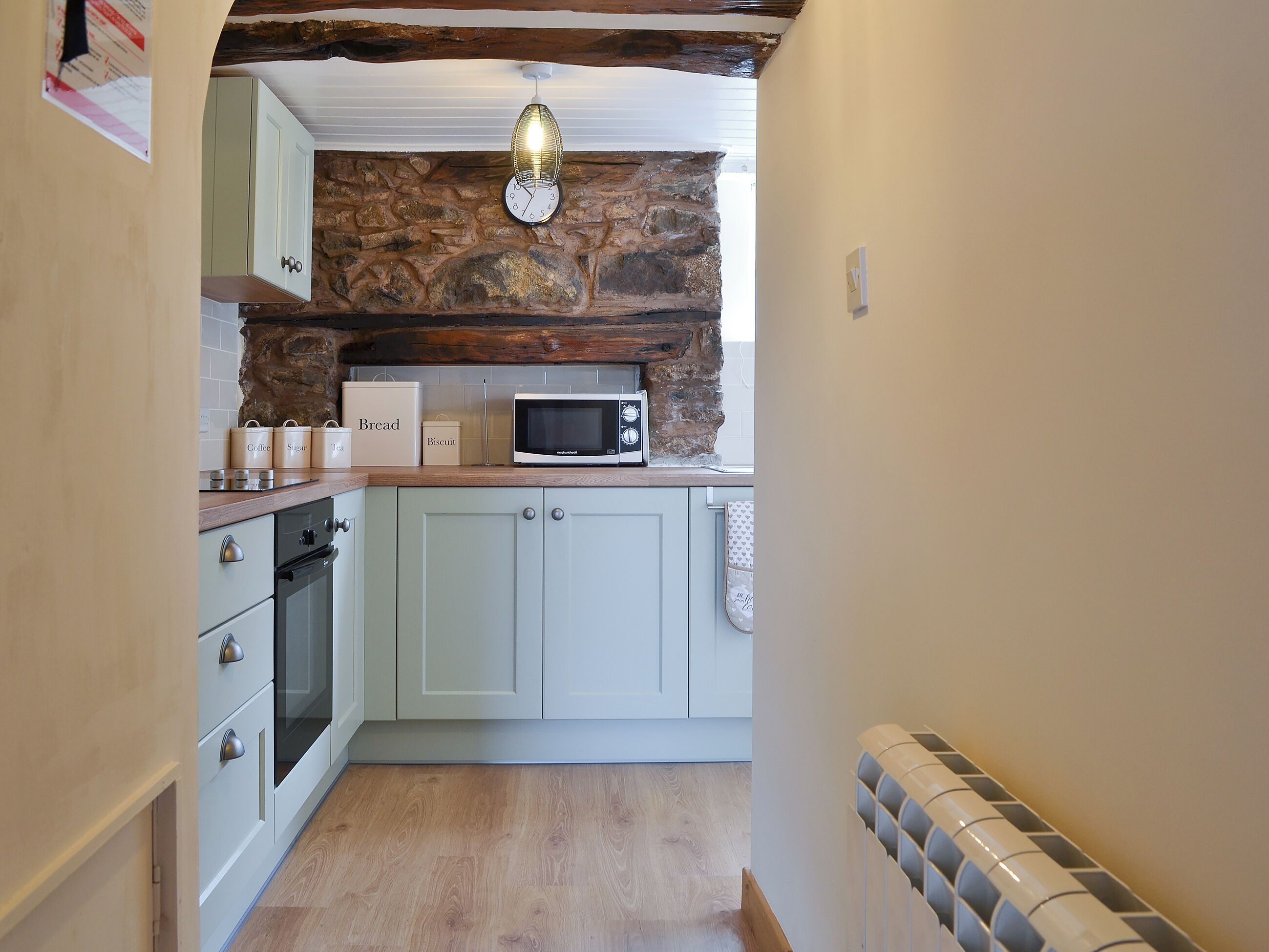 A narrow kitchen with stone walls, a microwave, and storage containers labeled 'Bread,' 'Coffee,' 'Sugar,' and 'Tea.'