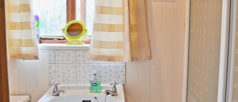 Um banheiro com pia, duas torneiras, uma janela com cortinas, um espelho e vaso sanitário.