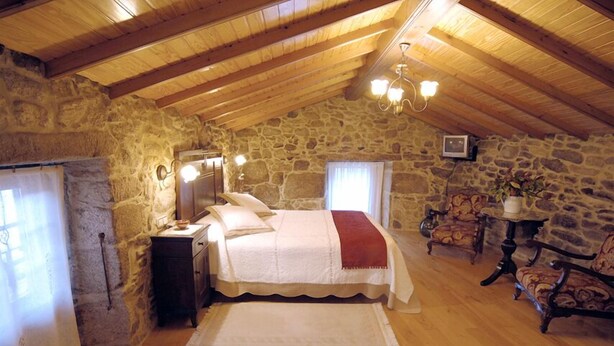 A cozy bedroom with a stone wall, wooden ceiling beams, a bed with a white and red blanket, a nightstand, a chair, and a small table with a lamp. - Os Migueliños (Catoira)