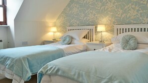 Un dormitorio con dos camas, cada una con una mesita de noche y una lámpara. La habitación tiene techo inclinado y papel tapiz con motivos florales.