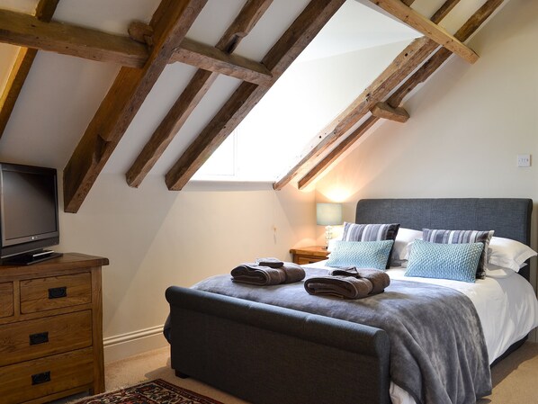 Une chambre avec un plafond en pente, un lit avec des oreillers, une table de chevet avec une lampe et un téléviseur posé sur une commode en bois.