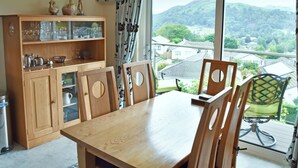 A dining area with a wooden table and chairs, a wooden cabinet with glassware, and a view of a residential area through large windows. - Badgers Rake 12 Squirrels Nest (Ambleside)