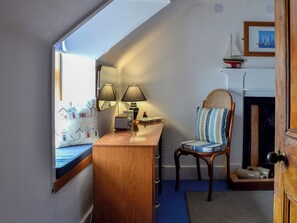 Een kamer met een houten bureau, twee lampen, een stoel met een gestreept kussen, een klein model schip en een lijst met een afbeelding aan de muur.