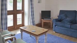Ein Wohnzimmer mit Couchtisch aus Holz, einem blauen Sofa, einem Fernseher auf einem kleinen Tisch und Blick durch ein großes Fenster mit Vorhängen.