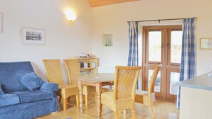 Ein gemütliches Wohnzimmer mit einem blauen Sofa, Korbstühlen, einem Holzesstisch und einer Küchenzeile mit Fenster und Vorhängen.