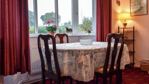 A dining area with a table covered in lace, two chairs, a window with curtains, a radiator, a lamp, and a wall-mounted shelf with a decorative item.