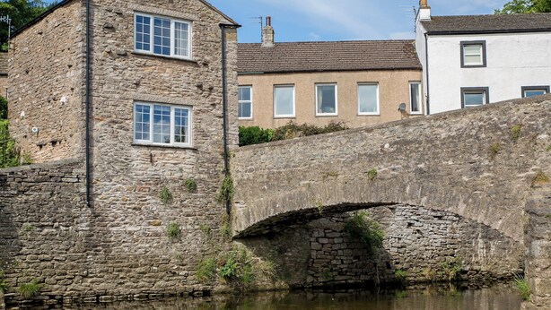 A stone bridge over a calm river, with buildings on either side, including a two-story stone house and a white building with multiple windows.