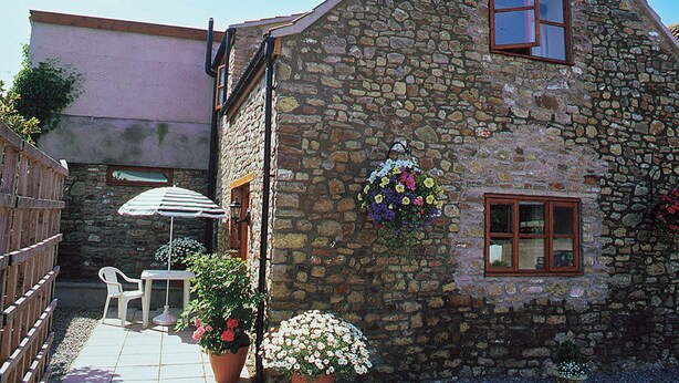 Ein Steinhaus mit kleiner Terrasse, Holzlattenzaun und Blumentöpfen.