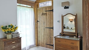 A bedroom with a wooden door, a dresser, a mirror, a bedside table with a vase of sunflowers, and a bed with floral bedding. - Romantic hideaway for couples (and their pets. (Carlton, near Saxmundham)