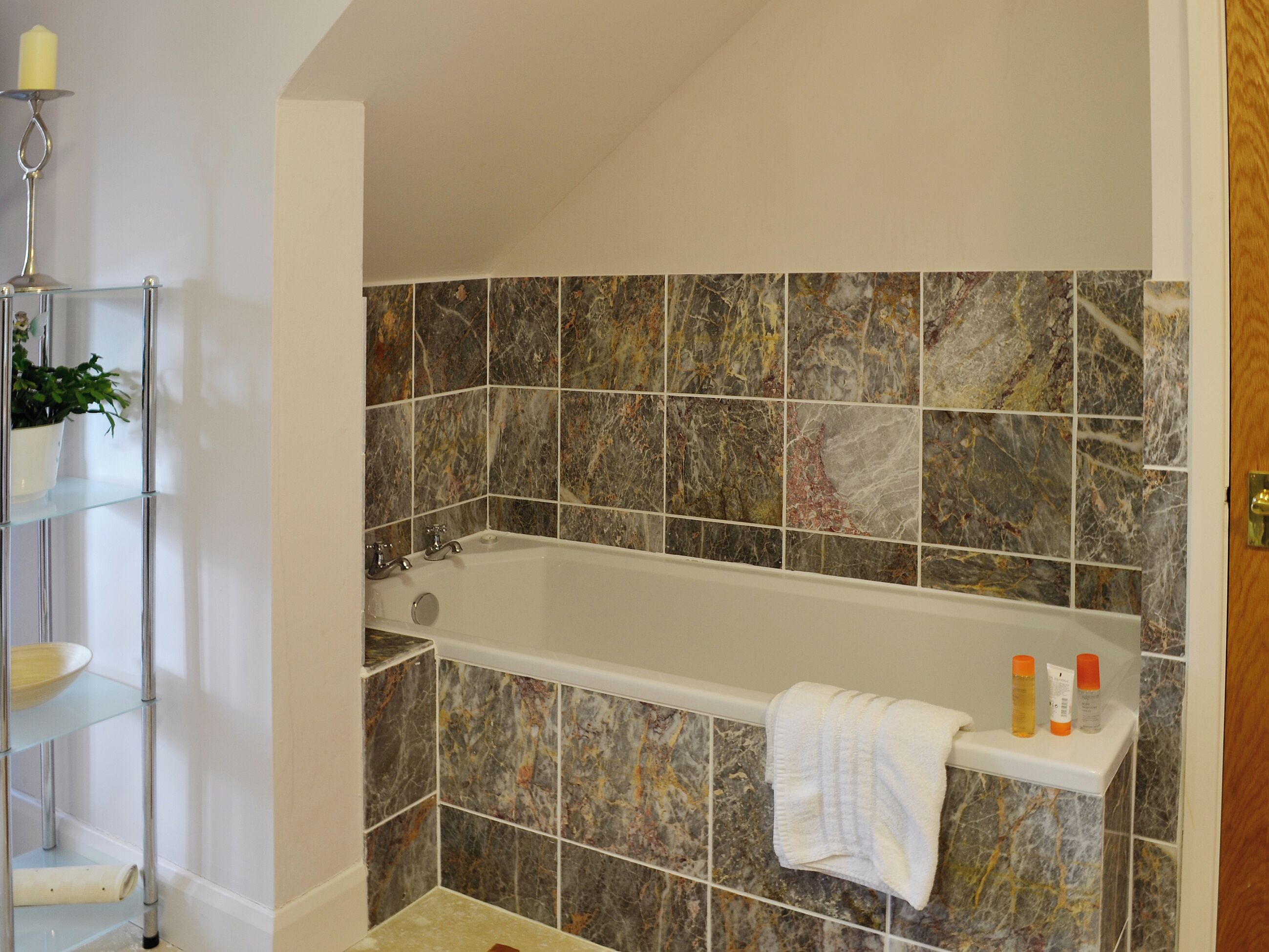 A bathroom with a bathtub, marble tiles, a shelf with toiletries, and a potted plant.