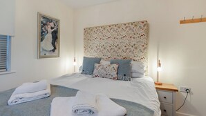 A neatly made bed with white linens and a grey throw, a bedside lamp, a nightstand with a lamp, and a floral-patterned headboard. - Short breaks available all year. (Keswick)