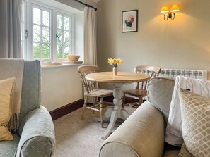 A cozy room with a round wooden table, two chairs, a sofa, a window with curtains, a wall-mounted light fixture, and a framed picture.