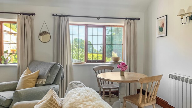 Una habitación acogedora con sofá, una mesa redonda con sillas, ventana con cortinas, radiador y una lámpara de pared.