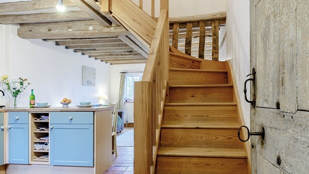 Un escalier en bois, doté d’une finition rustique, mène à une pièce comprenant un coin cuisine avec des armoires bleues et un sol carrelé.