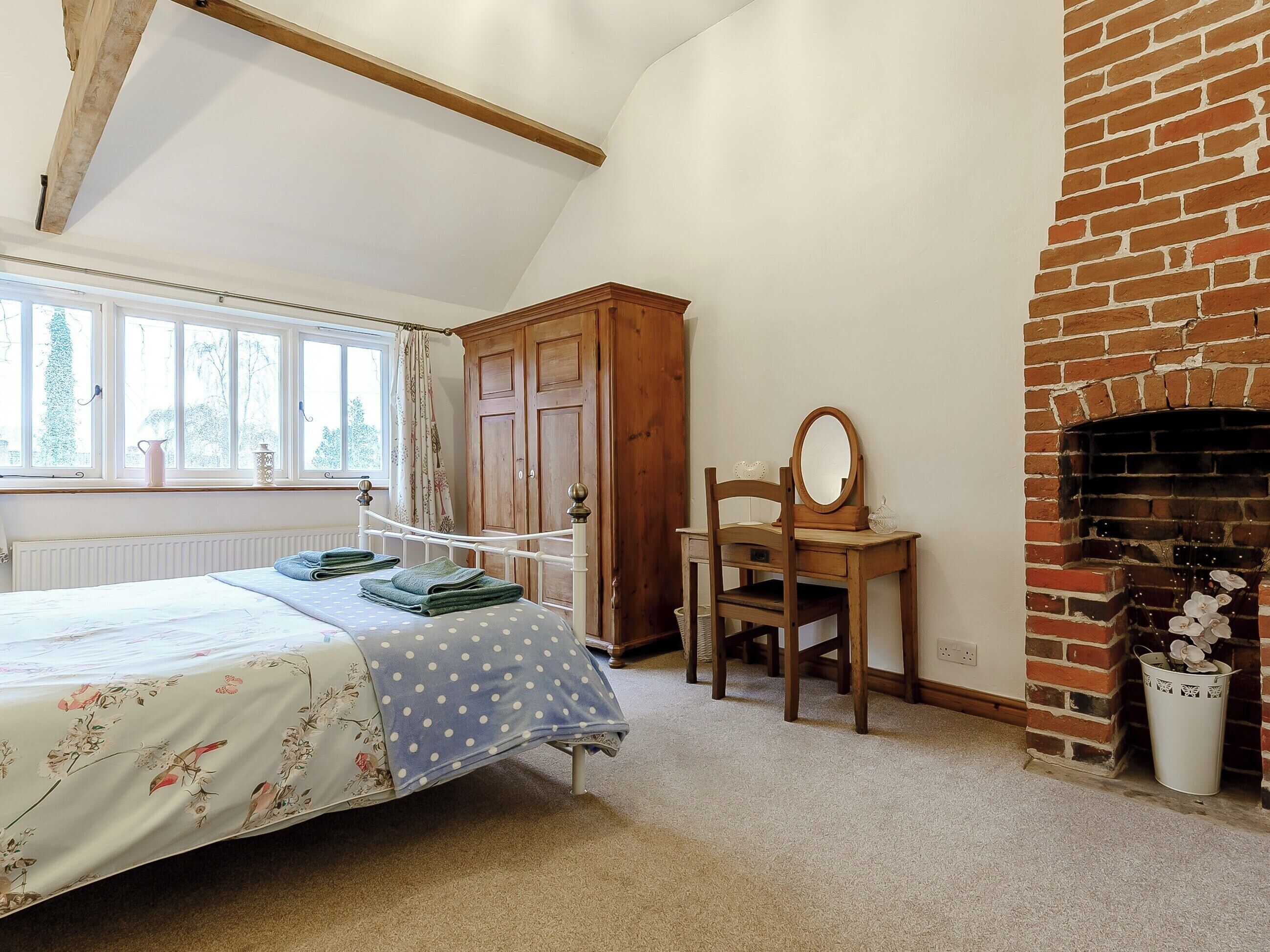 A bedroom with a bed, a wooden dresser, a chair with a mirror, a fireplace, and a window with curtains.