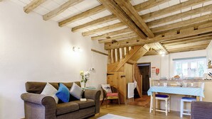 Un salon chaleureux avec un plafond en bois, un canapé agrémenté de coussins à motifs, une table à manger recouverte d’une nappe bleue et, au fond, un coin cuisine.