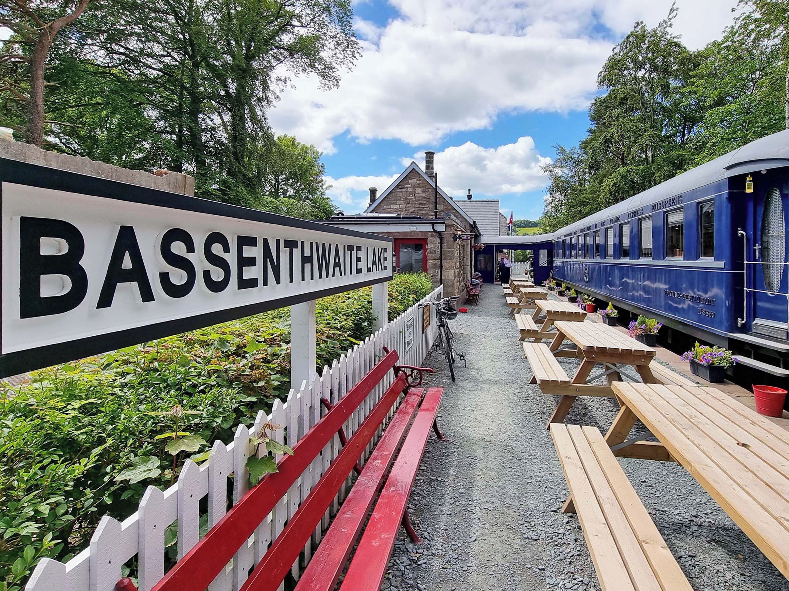 A sign for BASSENTHWAIT LAKE, a wooden bench-lined pathway, and a blue train with a dining car.