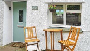 A cottage with a green door, a sign reading 'OSPREY COTTAGE,' two wooden chairs, a small table, and a hanging flower basket. - 2 bedroom accommodation in Bassenthwaite, near Keswick (Bassenthwaite, near Keswick)