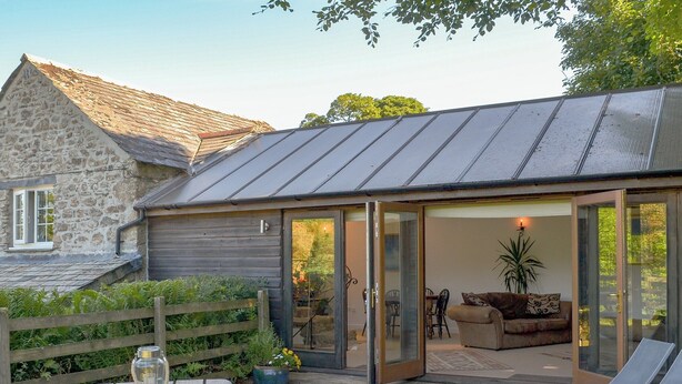 Une terrasse en bois avec une table de pique-nique et des chaises longues, un bâtiment en pierre avec un toit en pente, et une porte vitrée donnant sur un espace intérieur aménagé avec un canapé et un pot de fleurs.