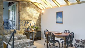Une pièce chaleureuse avec un mur en pierre, un plafond en bois et une lucarne. On y trouve une table à manger avec des chaises, ainsi qu’un petit escalier menant à une autre pièce.