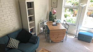 A living room with a blue sofa, a wooden coffee table, a dining table with chairs, a bookshelf, and a window with a view of greenery.