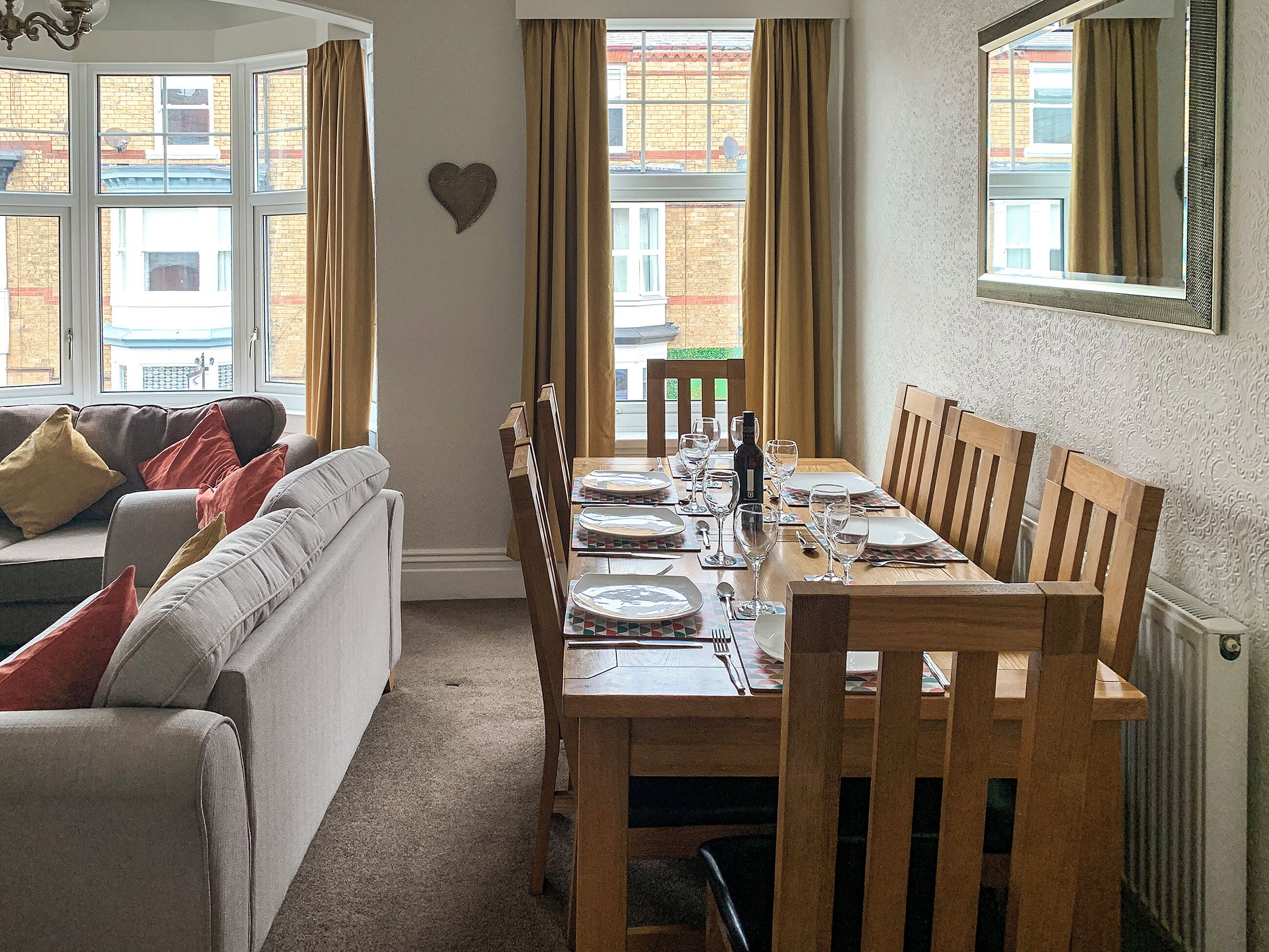 A dining area with a wooden table set for four, a sofa with cushions, and a heart-shaped decoration on the wall.