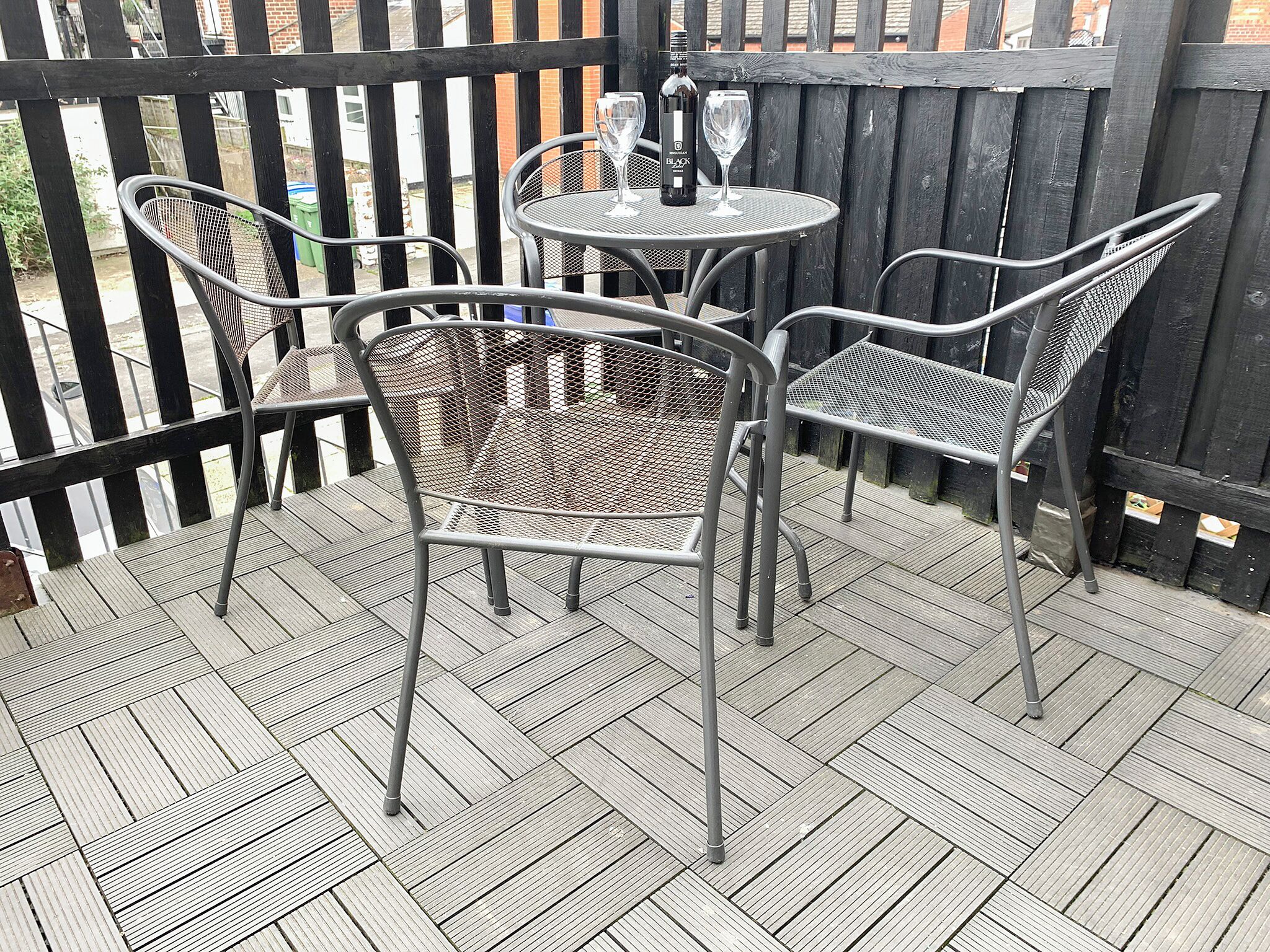 Un patio con sillas y una mesa de metal, una botella de vino Black y dos copas de vino.