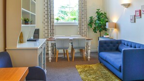 A living room with a blue sofa, a dining table with chairs, a small cabinet, and a window with curtains. - County House Apartment - 27911 (York)