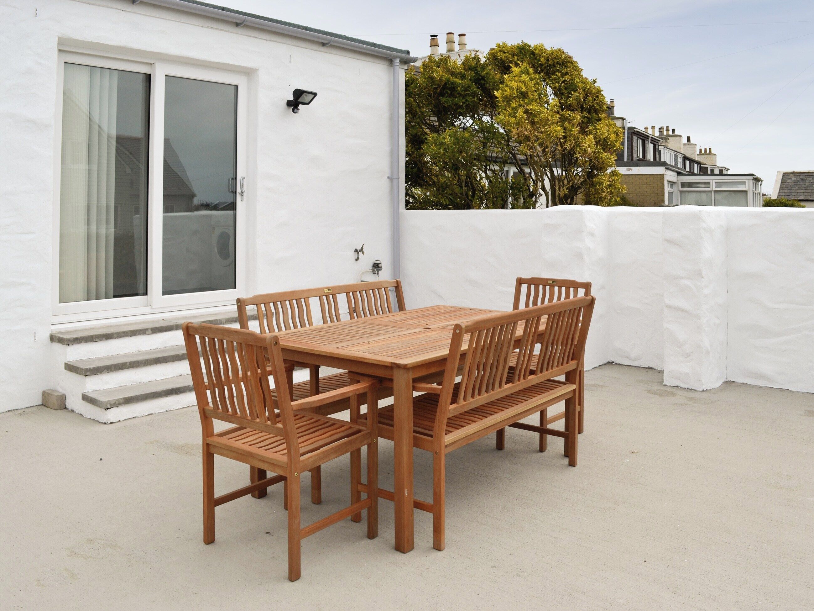 A wooden table and chairs set on a concrete patio with a white wall and a window in the background.