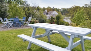 Une table de pique-nique avec des bancs, située dans un espace verdoyant, avec des chaises et une maison en arrière-plan.