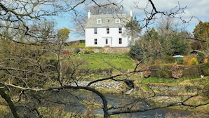 Un bâtiment blanc doté de nombreuses fenêtres, entouré d’arbres et bordé par un plan d’eau.