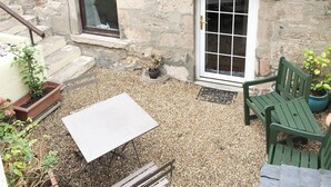 Una cabaña de piedra con un patio adoquinado, una mesa blanca y dos sillas verdes.