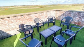 Un área con mesas y sillas de plástico al aire libre, rodeada por un muro de ladrillo y vista a un campo verde.