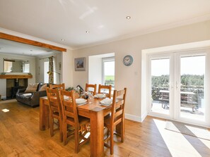 A dining area with a wooden table and chairs, a fireplace, and a view of the outdoors through large windows. - 2 bedroom accommodation in Kirkcudbright (Kirkcudbright)