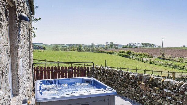 A hot tub with steps, set against a stone wall and a wooden fence, overlooking a green landscape.