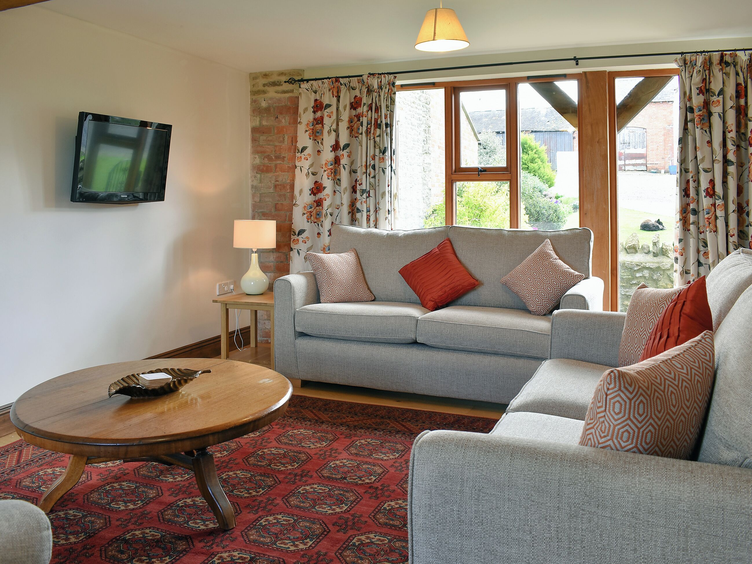 A living room with a grey sofa, patterned cushions, a wooden coffee table, a lamp, and a TV on the wall.
