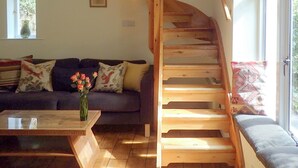 Un escalier en bois menant à un niveau supérieur, un canapé de couleur sombre agrémenté de coussins à motifs et une petite table sur laquelle est posé un vase de fleurs.