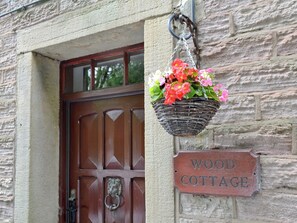 Una porta de fusta amb una làmina de bronze i una cistella penjant plena de flors roses. Hi ha un rètol que diu "WOOD COTTAGE".