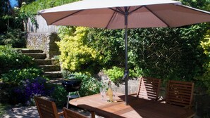 Ein Holztisch mit vier Stühlen und einem großen Sonnenschirm, platziert in einem Garten mit Steinstufen und üppigem Grün.