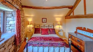 A bedroom with a stone wall, wooden beams, a bed with red bedding, two bedside tables with lamps, and a dresser. - 2 bedroom accommodation in Newton Reigny (Newton Reigny)
